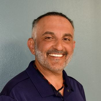 Photo of Dr. Afsheen smiling, wearing blue colored shirt and black and red beaded necklace against grey-blue wall background.
