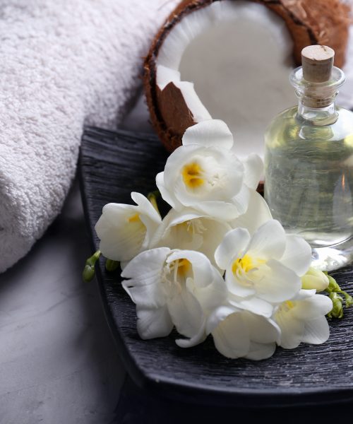 Chiropractic Accessories. White rolled up towel, cracked open coconut, and a black textured tray with white flowers, a bottle of oil and two lit candles.