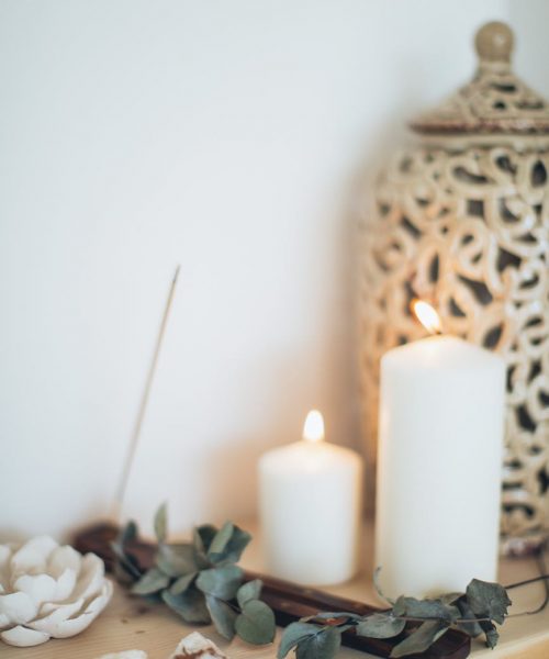 Spa counter with white lit candles, green leaves, sea shells, and a large patterned vase with white background.