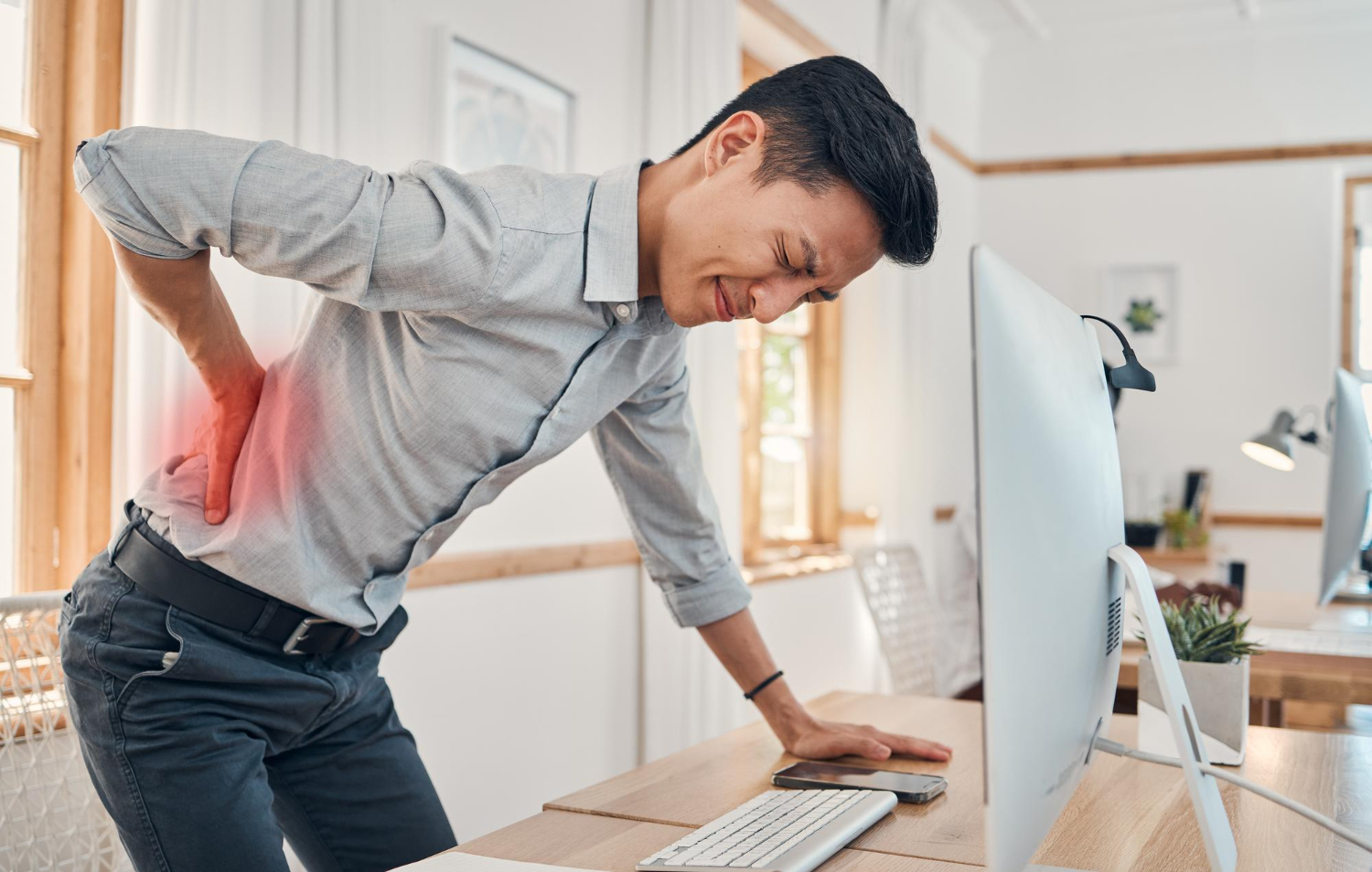 Man standing at desk, holding his lower back in pain, wincing while working at a computer.
