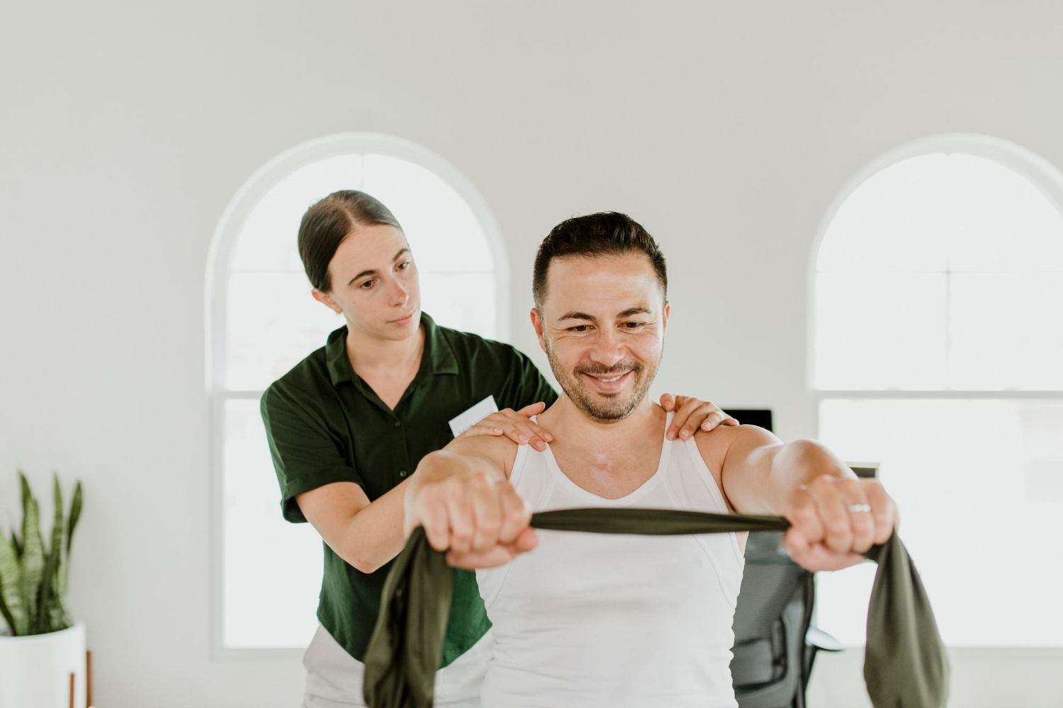 Smiling man does resistance band exercise with a therapist assisting him in a bright room.
