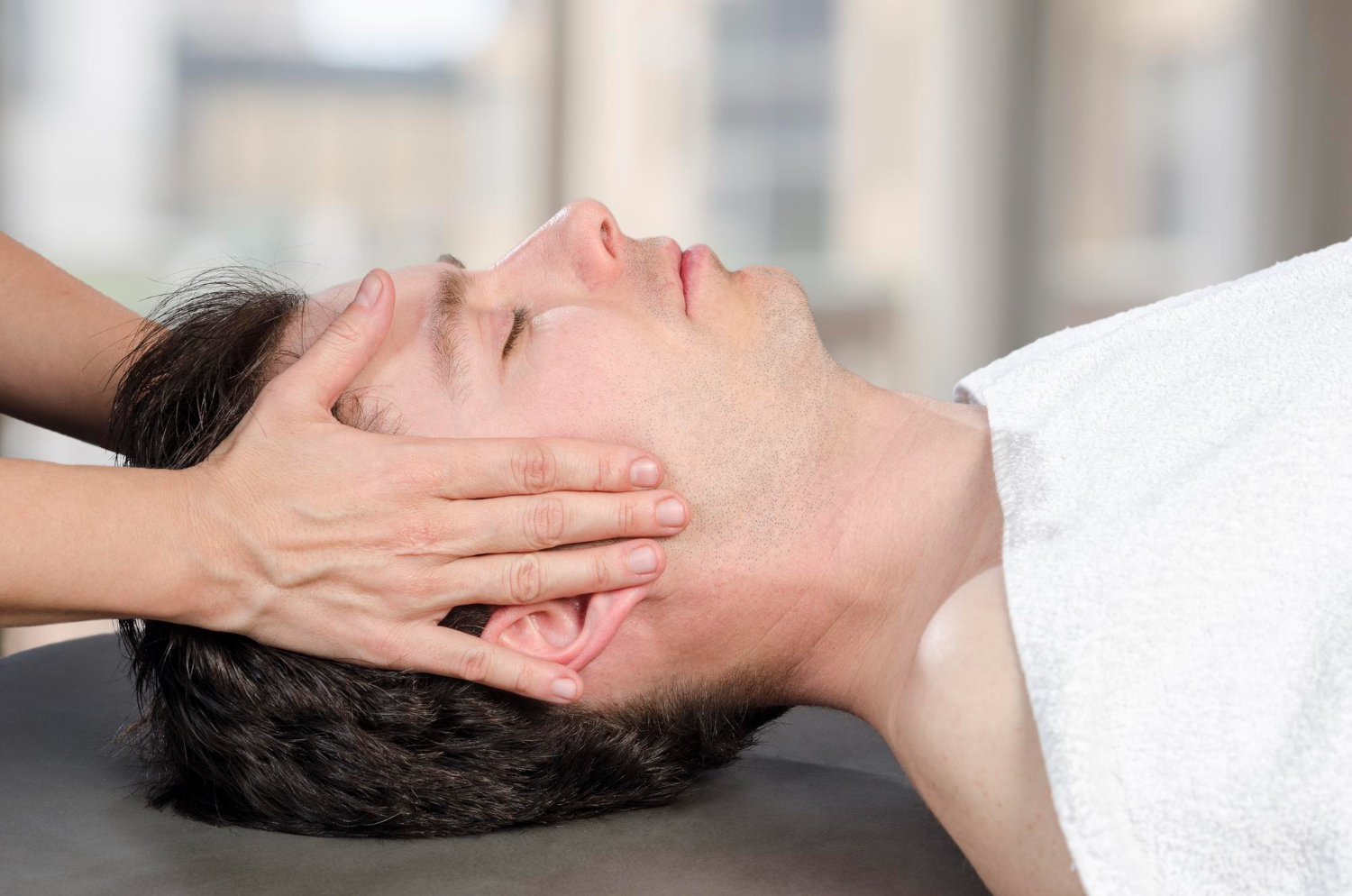 Person lying down receiving a head massage, covered with a white towel in a calming environment.