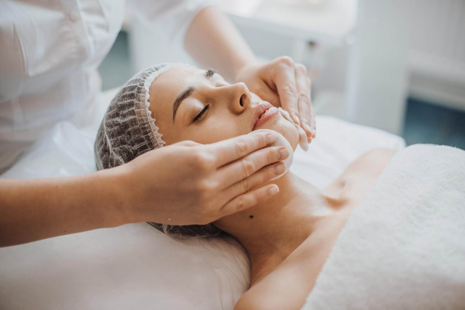A person receiving a facial treatment, lying down with eyes closed and a hair net, as hands massage their face.